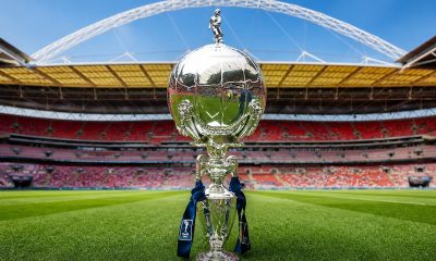 FA Trophy cup displayed on a stadium pitch with stands and arch in the background