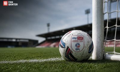 A Sky Bet League Two match ball resting against a white goalpost on a grass pitch with an empty stand in the background.