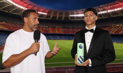 A pundit and a teenage footballer stand inside a near-empty stadium; the teenager holds a Premier League‑branded prop while the presenter speaks into a microphone.