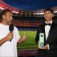 A pundit and a teenage footballer stand inside a near-empty stadium; the teenager holds a Premier League‑branded prop while the presenter speaks into a microphone.