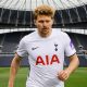 A footballer in a white Tottenham Hotspur jersey running on the pitch inside an empty stadium.